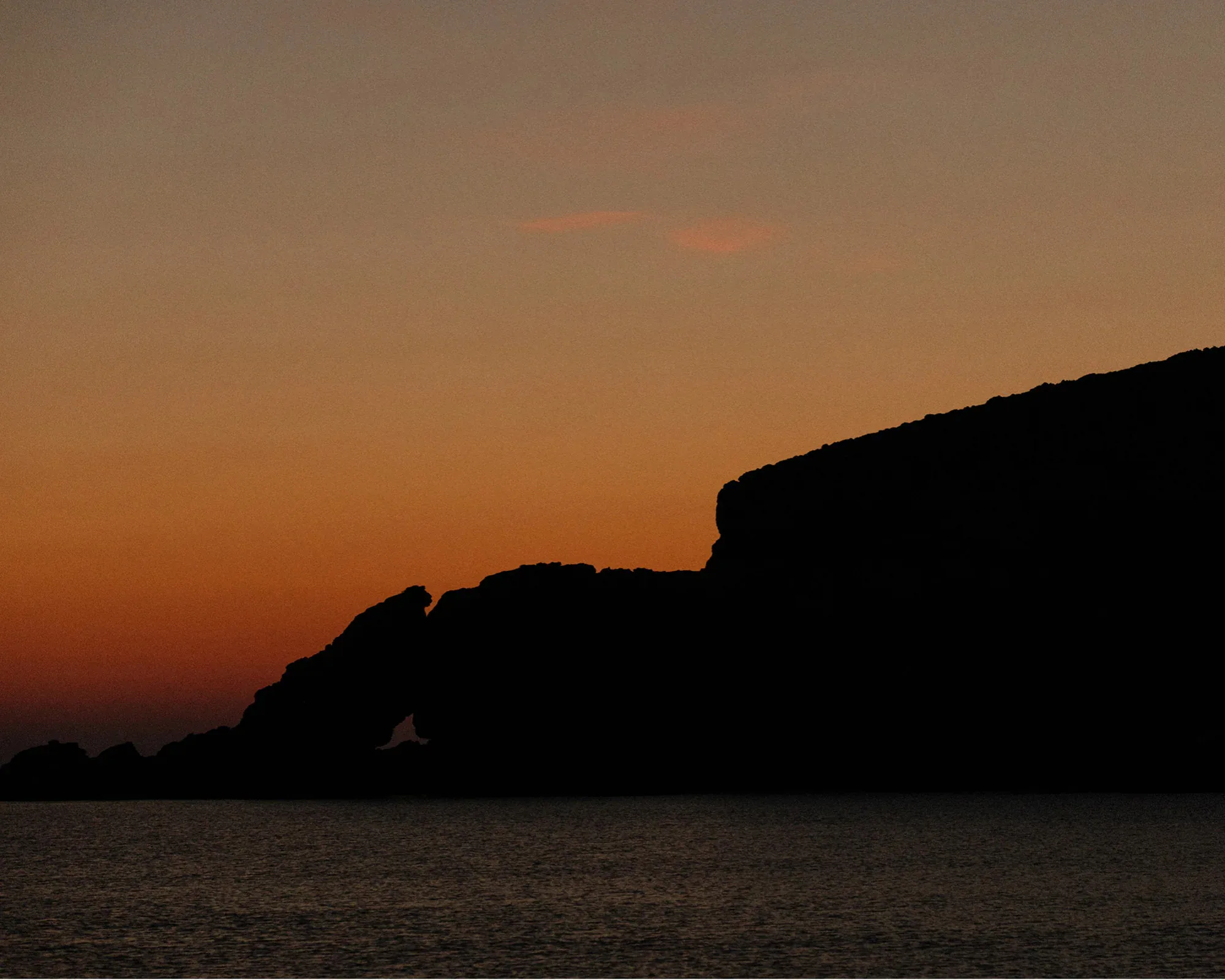 The sun setting behind a headland on Mykonos, an orange sky above a dark silhouetted coastline and still sea.